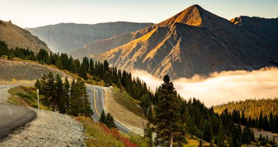 A landscape scene at sunset, with a blue sky behind sloped mountains, low clouds rolling though a valley, a forest, and a curvy mountain road