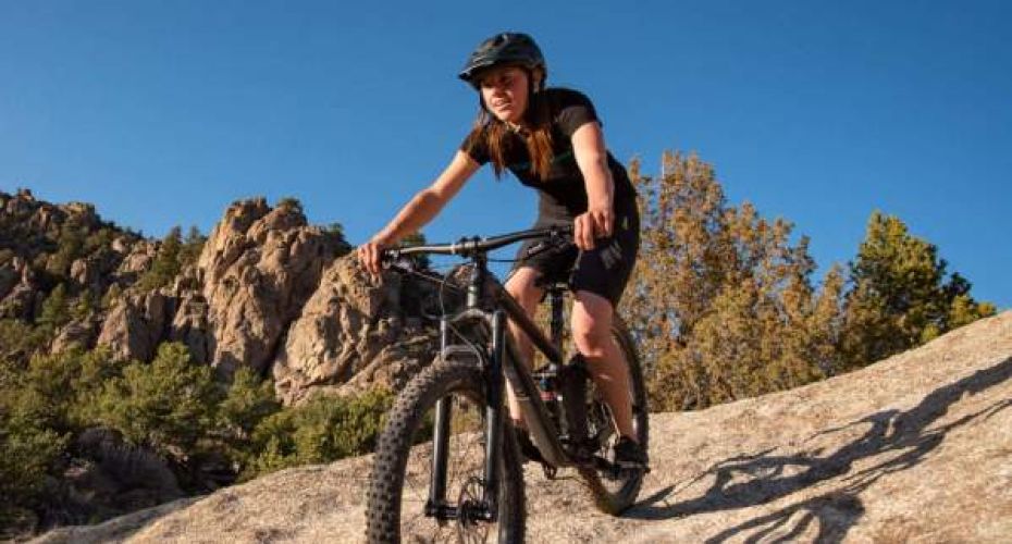 A woman mountain biking down a rocky path, with a forested, rocky hill and blue skies behind her