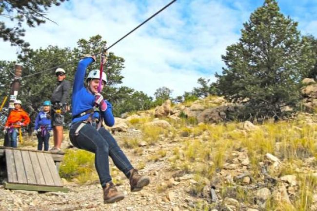 A person zip lining against a cloudy blue sky and hills, with several people behind them waiting for their turn