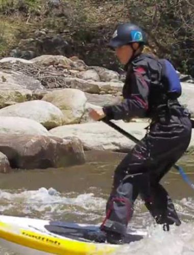 A man paddle boarding down a whitewater rapid, standing up with a paddle behind him